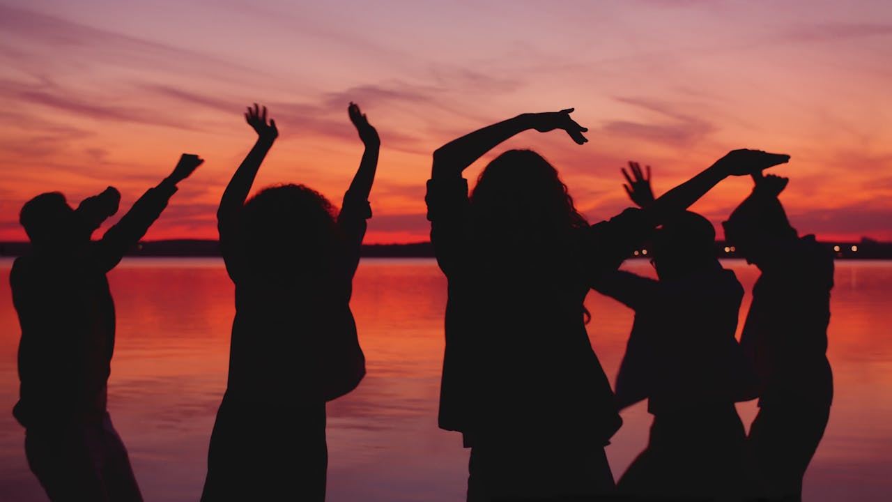 Silhouetted group of friends dancing joyfully by a tranquil lake during sunset.